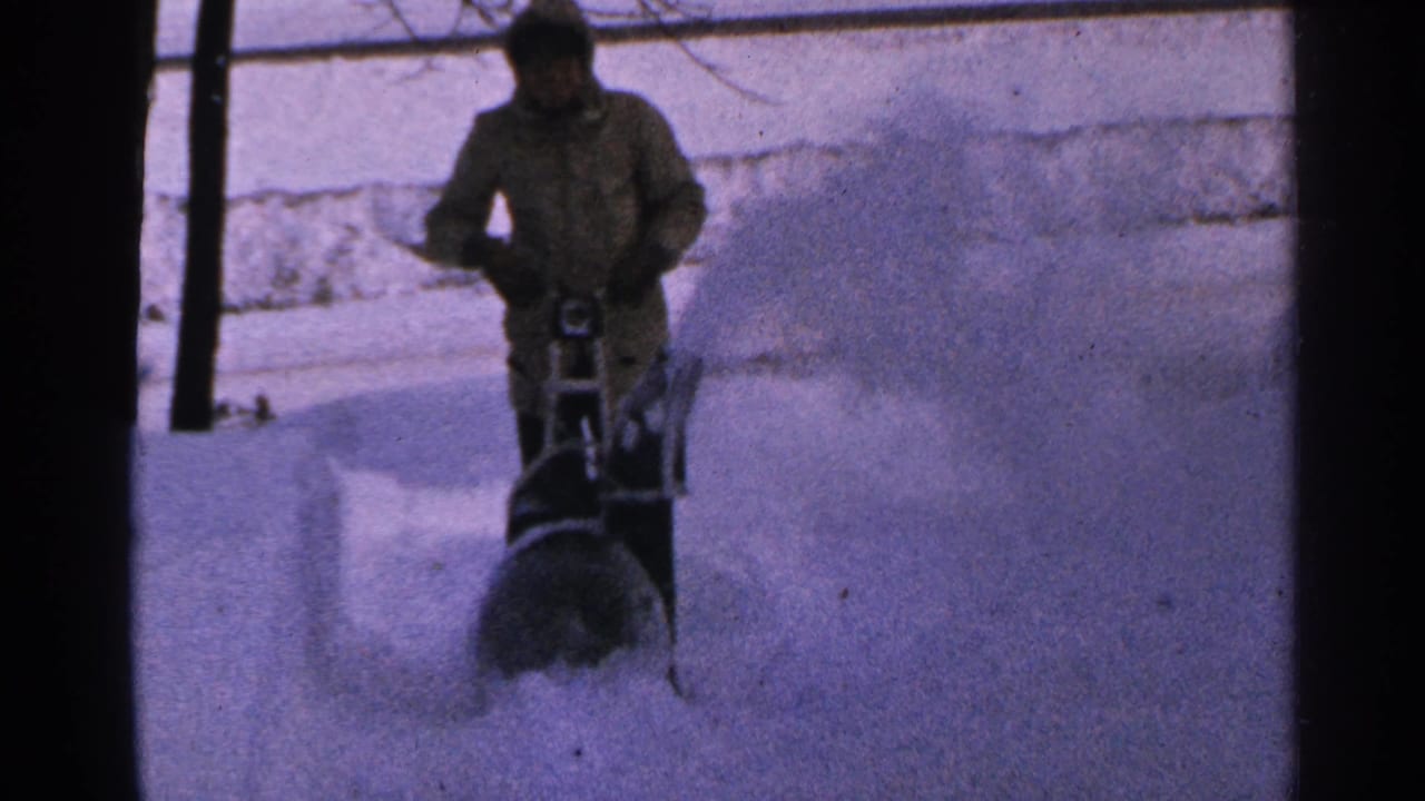 In 1965 Minnesota home-movie footage, a bundled person operates a snow blower through deep snow beside bare trees.
