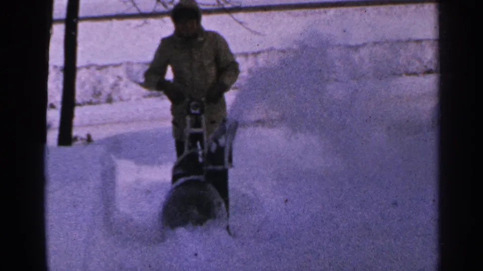 In 1965 Minnesota home-movie footage, a bundled person operates a snow blower through deep snow beside bare trees.