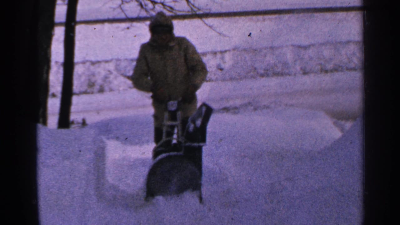 A bundled person in Minnesota operates a walk-behind snow blower, sending powder across a snow-covered driveway.