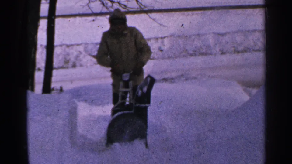 A bundled person in Minnesota operates a walk-behind snow blower, sending powder across a snow-covered driveway.