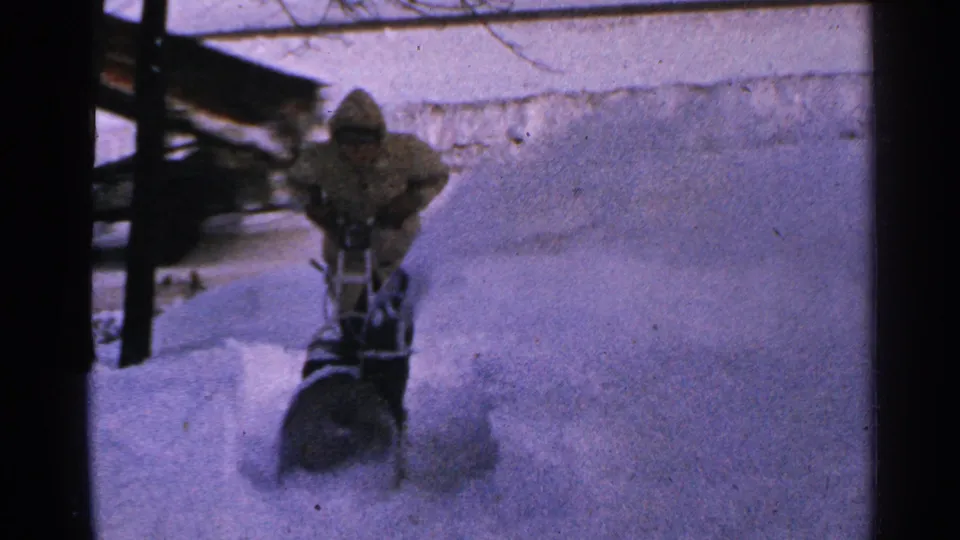 A bundled operator continues snow blowing in 1965 Minnesota, with powder rising beside a dark roof or structure edge.