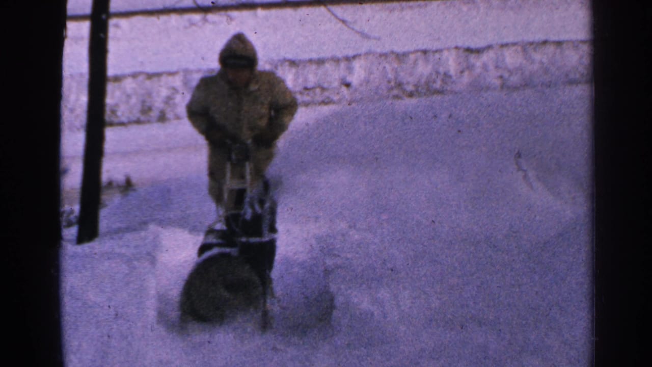In 1965 Minnesota home-movie footage, a bundled person operates a snowblower as snow sprays across a buried walkway.