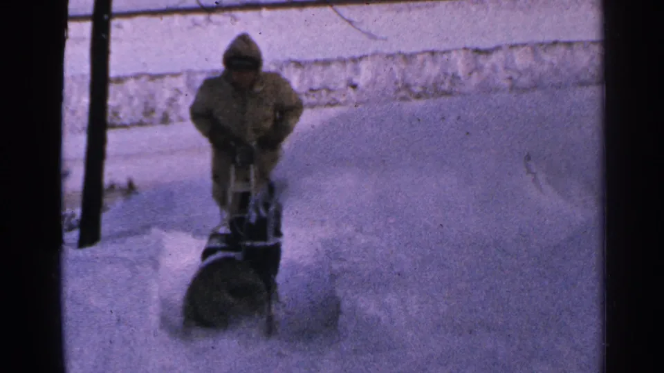 In 1965 Minnesota home-movie footage, a bundled person operates a snowblower as snow sprays across a buried walkway.