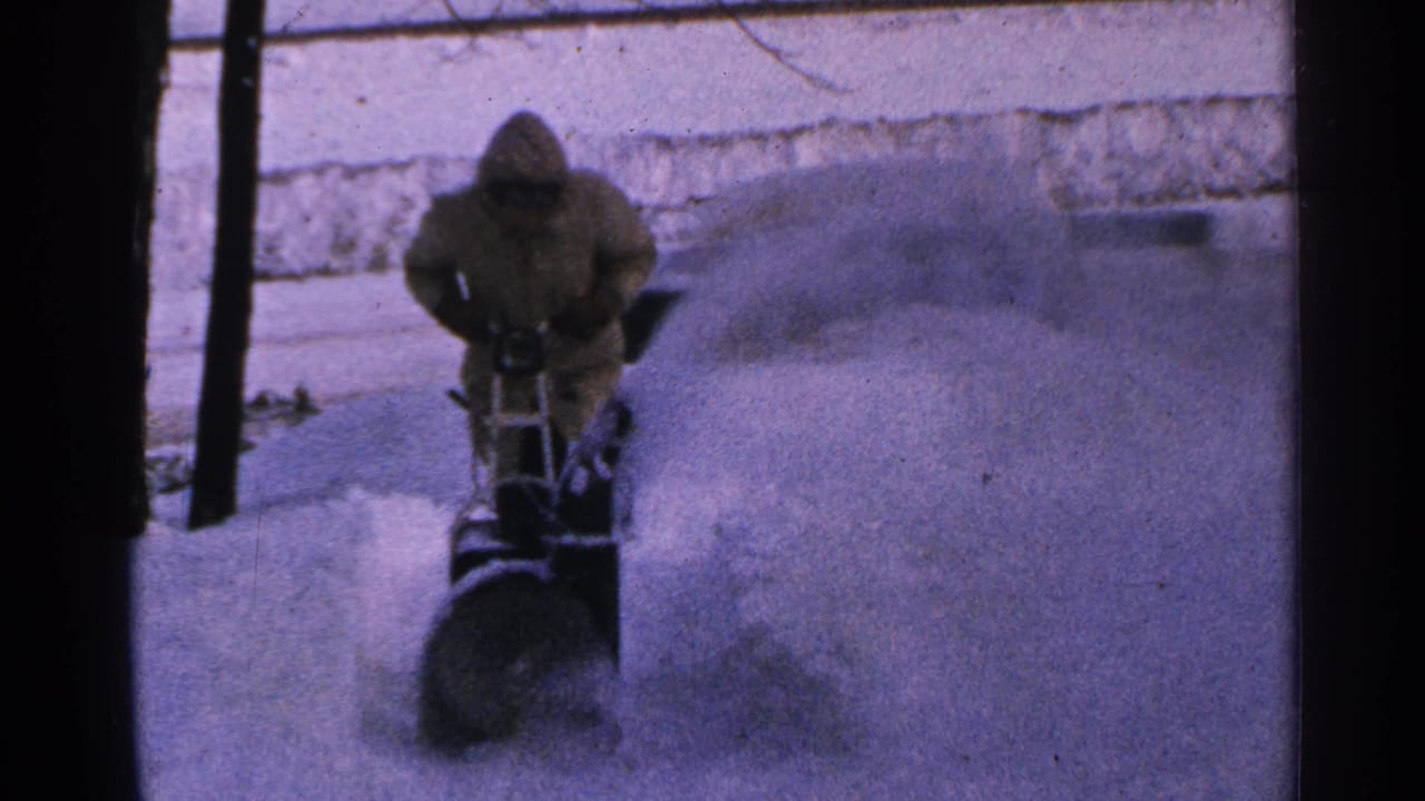 A hooded person in 1960s Minnesota footage guides a snowblower as a large plume arcs across the snowy ground.