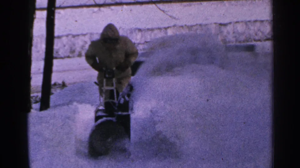 A hooded person in 1960s Minnesota footage guides a snowblower as a large plume arcs across the snowy ground.