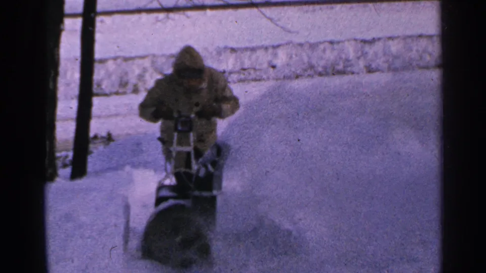 The bundled operator leans into the snowblower while clearing deep snow in a grainy 1965 Minnesota home-movie frame.