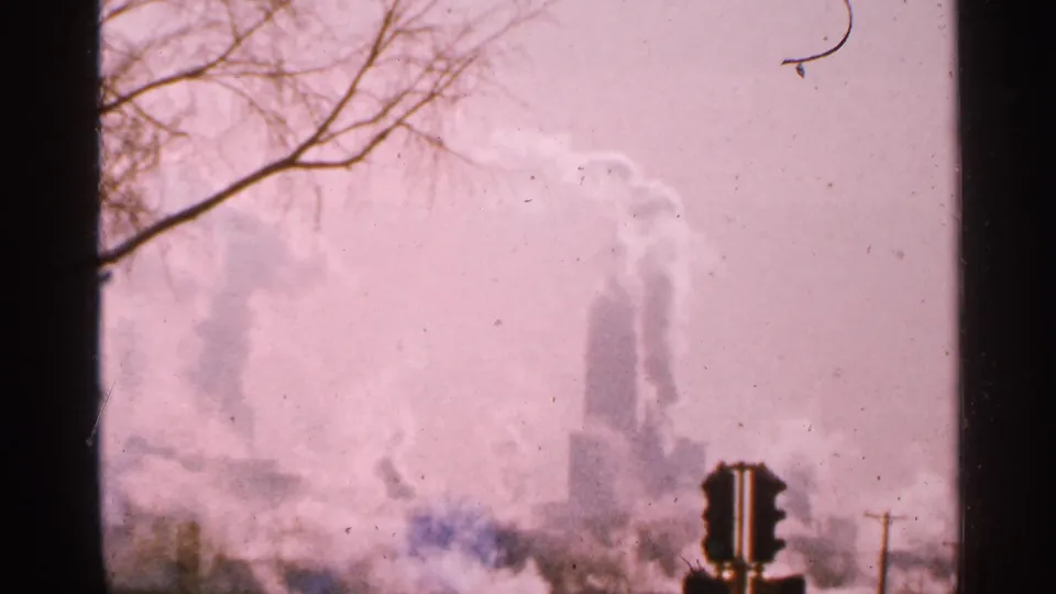 1965 Minnesota 8mm frame showing industrial smoke plumes, a traffic light, bare branches, and a pink haze over the city view.