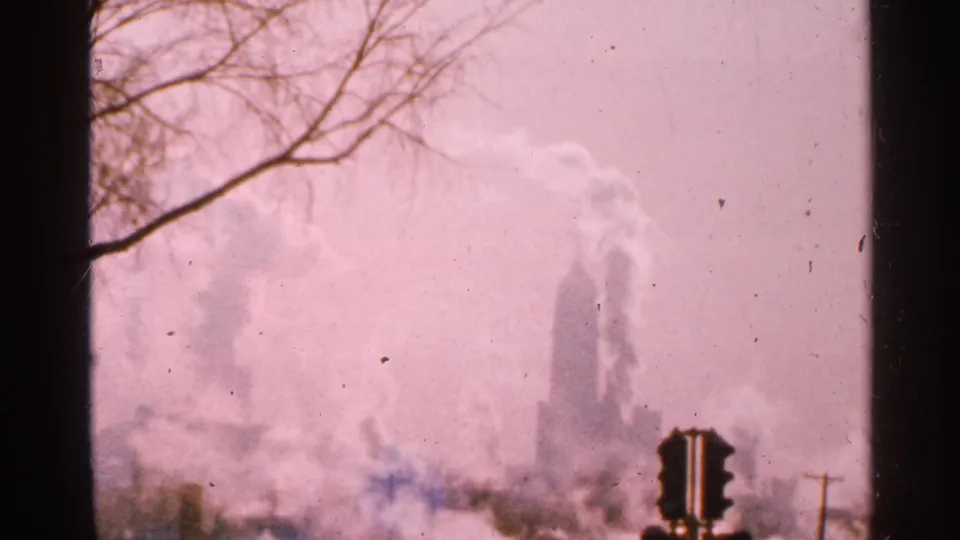 1965 Minnesota 8mm frame with tall smoke plumes, a silhouetted traffic light, utility poles, and a hazy industrial skyline.