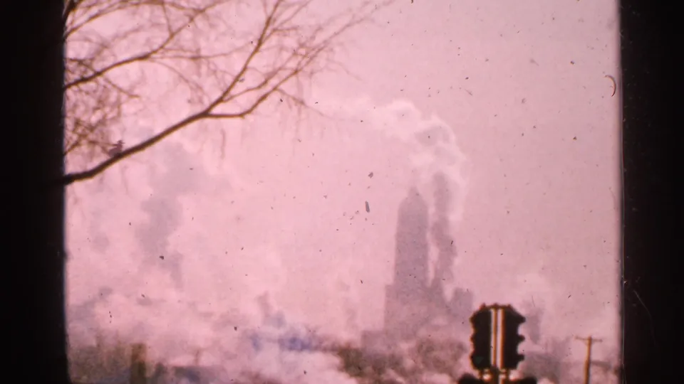 1965 Minnesota 8mm frame showing a large industrial smoke column, traffic signal, bare branches, and faded color haze.