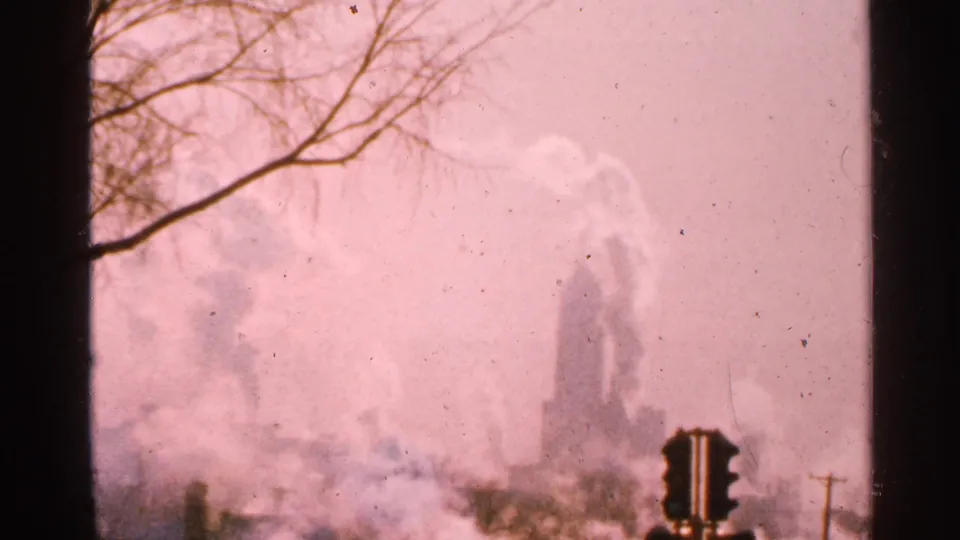 1965 Minnesota 8mm frame of industrial smoke plumes drifting through haze, with a foreground traffic light and bare roadside branches.