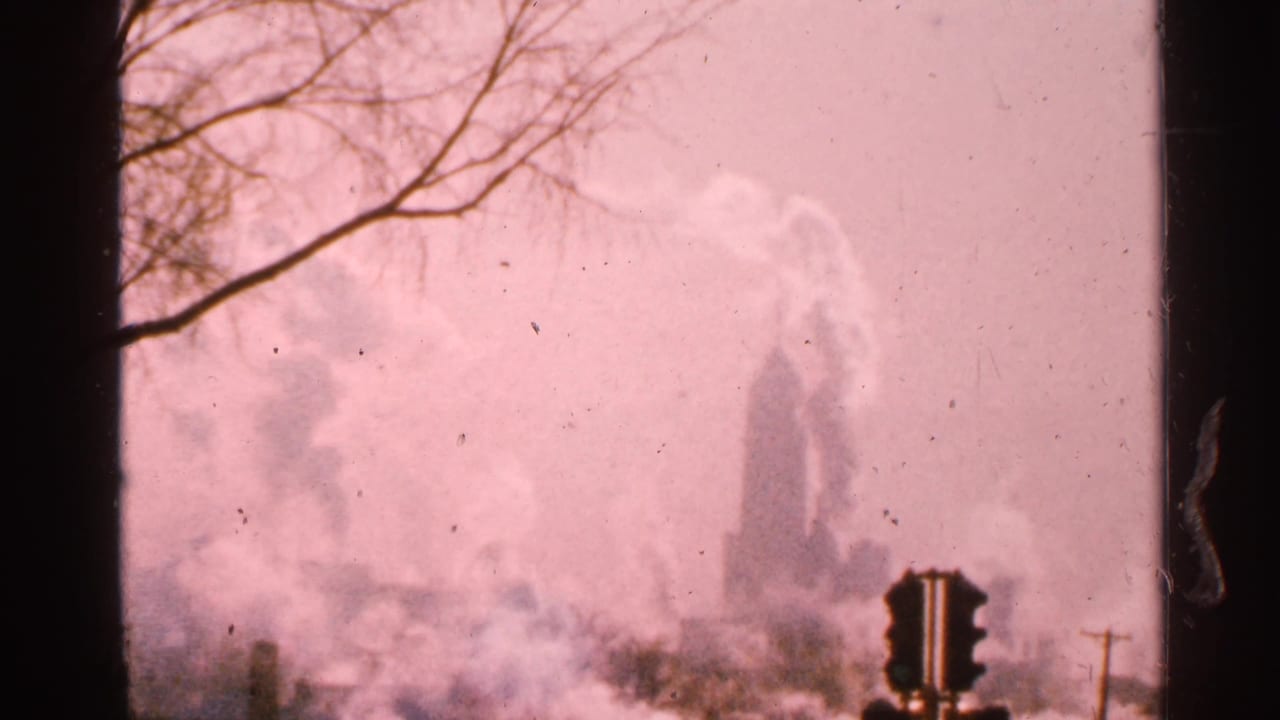 1965 Minnesota 8mm frame showing a hazy industrial skyline with smoke, bare tree branches, pale sky, and dark film borders.