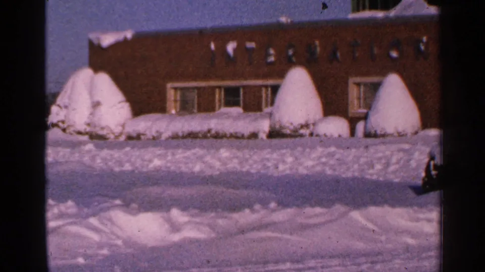 1965 Minnesota 8mm frame showing a closer view of the brick facade, partial INTERNATIONAL lettering, windows, and snowy shrubs.