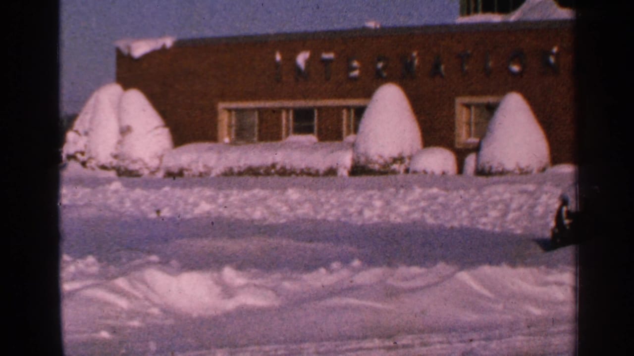 1965 Minnesota 8mm frame showing a brick institutional building, windows, and snow-laden shrubs behind a deep snowbank.
