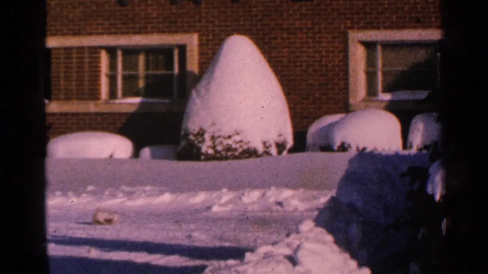 A large snow-capped shrub, brick wall, and window blinds appear in a 1965 Minnesota winter home-movie frame.