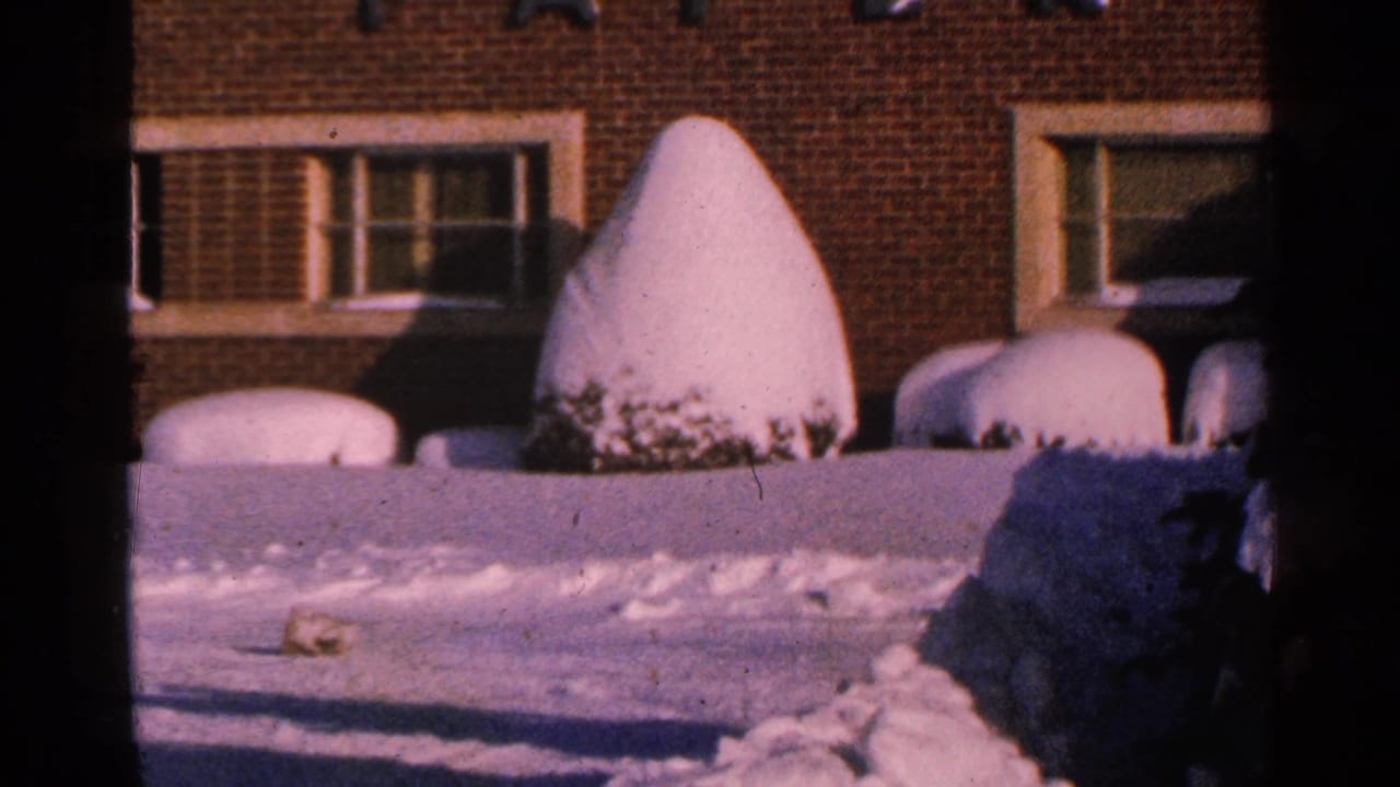 1965 Minnesota home-movie frame showing a brick residential exterior, deep snow, and a conical evergreen shrub under winter light.