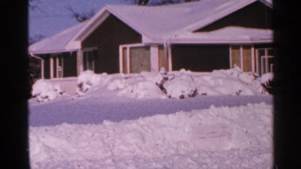 1965 Minnesota 8mm frame showing a snow-covered house, yard, shrubs, and dark tree trunks.