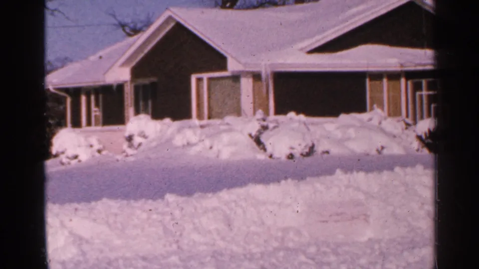 1965 Minnesota winter home-movie frame of a low house, snow-covered roof, yard, and shrubs.