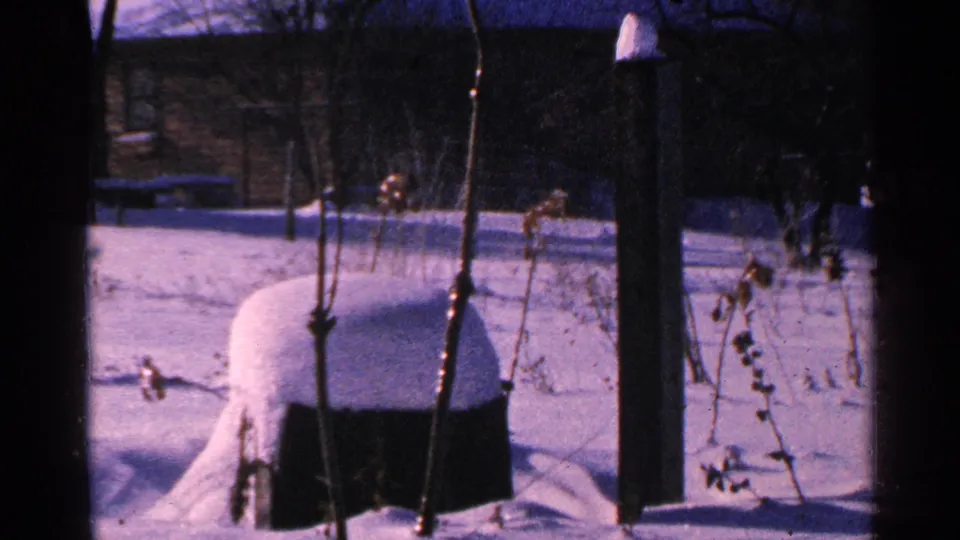 1965 Minnesota 8mm view of a snow-filled yard with a covered object and bare winter plants near porch posts.