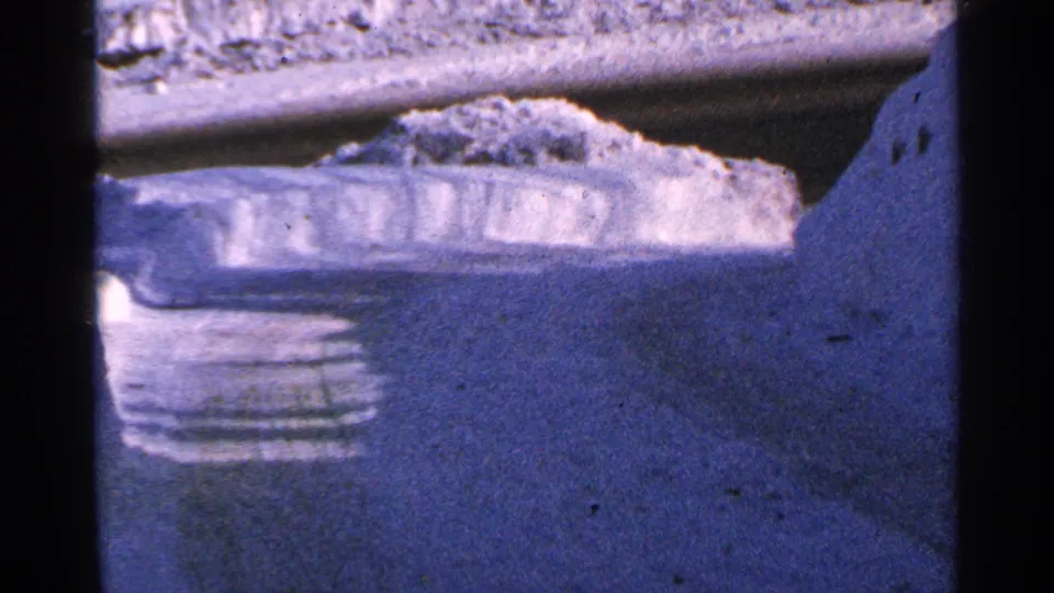 1965 Minnesota home-movie frame showing a steep snowy roadway edge with piled snow and a limited view around the corner.