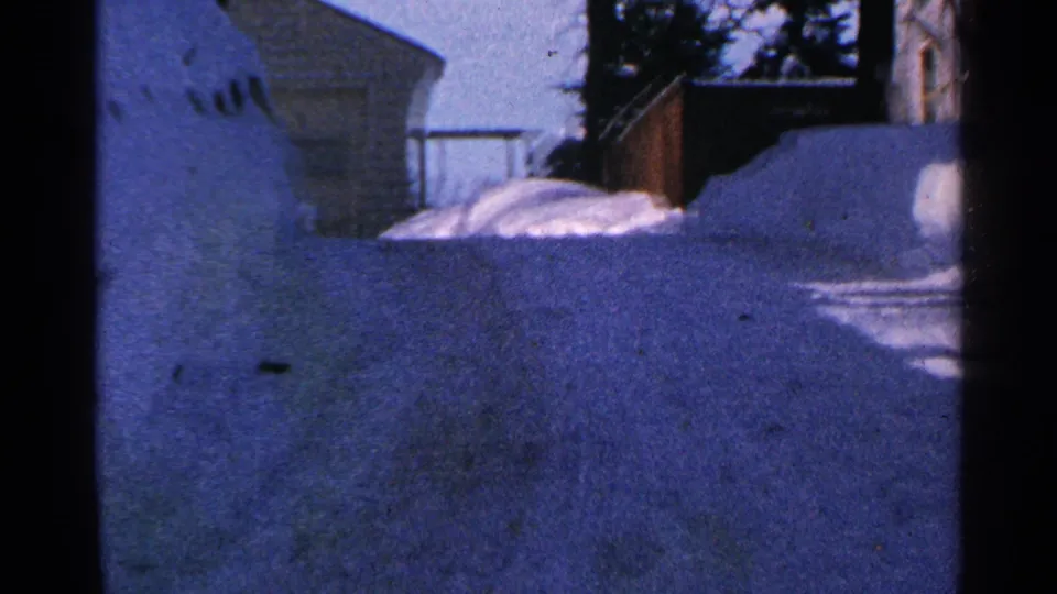 Wider 1965 Minnesota 8mm view of a snow-covered residential corner with a house edge, fence, trees, and deep banks.