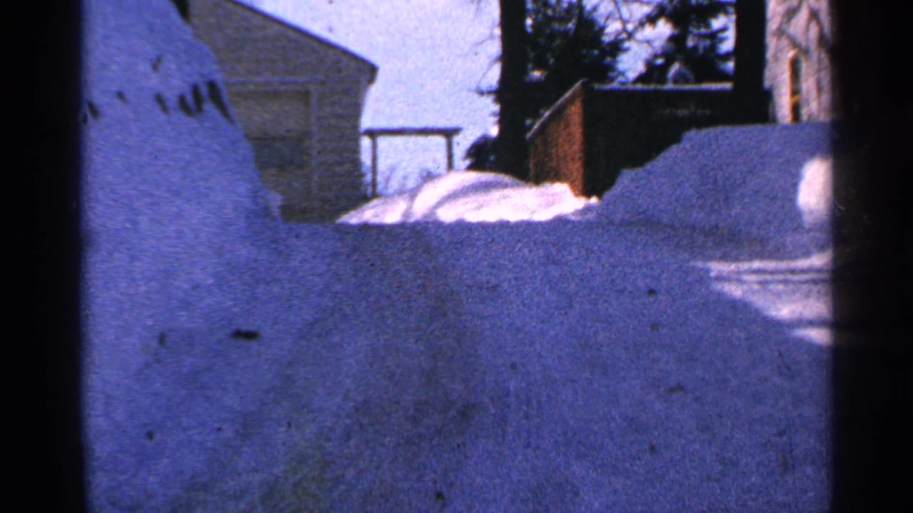 Snow-covered residential view with house roofline, trees, and high snowbanks.