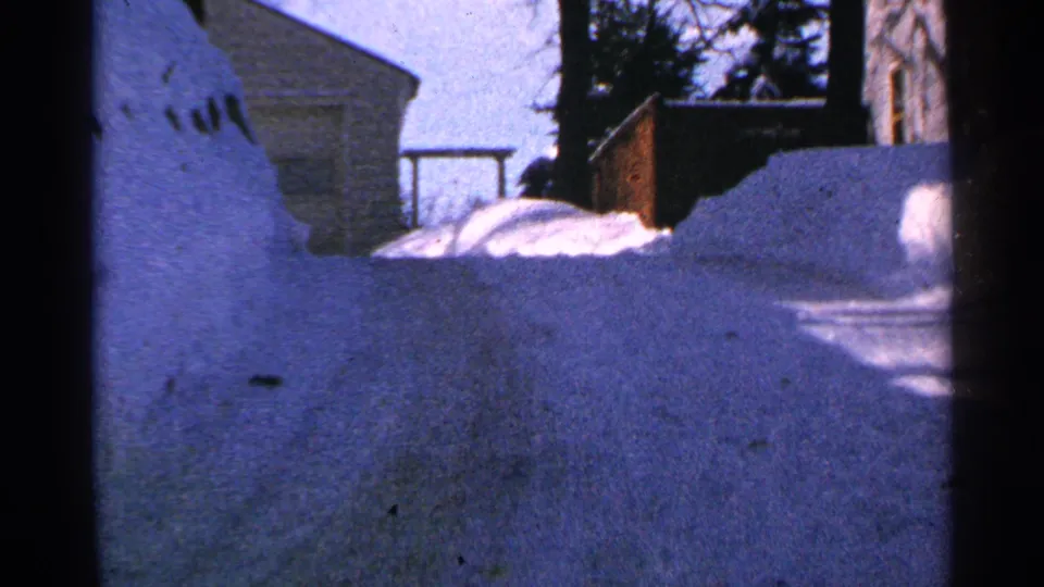 1965 Minnesota winter frame showing a low-angle view past deep snow toward a house, bare trees, and a small railing or fence.