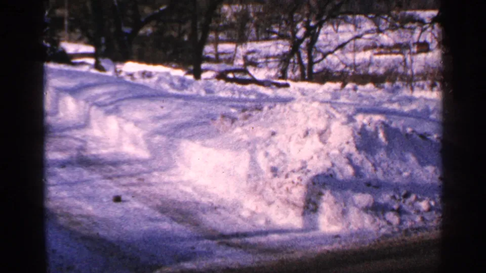 1965 Minnesota 8mm frame looking down a plowed residential street or driveway bordered by snowbanks, with trees and houses in the background.