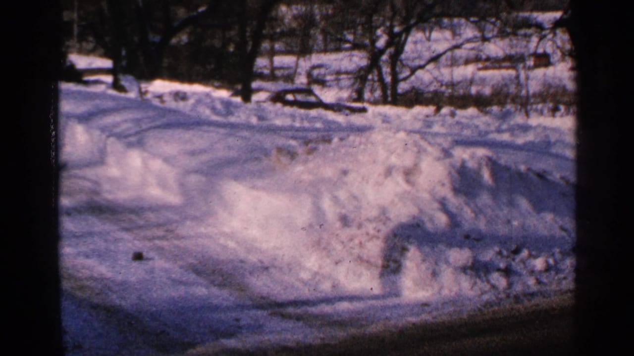 1965 Minnesota 8mm frame showing a plowed snowy roadside, high snowbanks, bare trees, and a small distant vehicle-like shape.