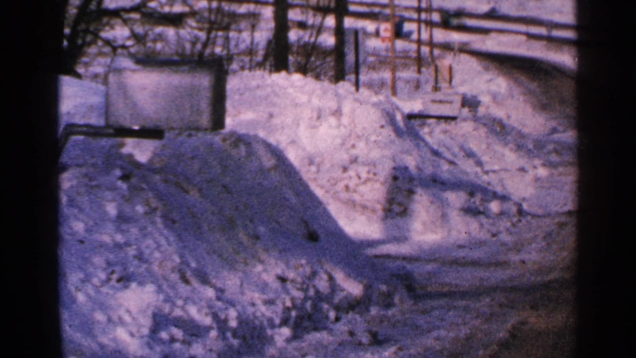 1965 Minnesota 8mm frame showing piled roadside snow, bare trees, utility lines, and a pale winter sky.