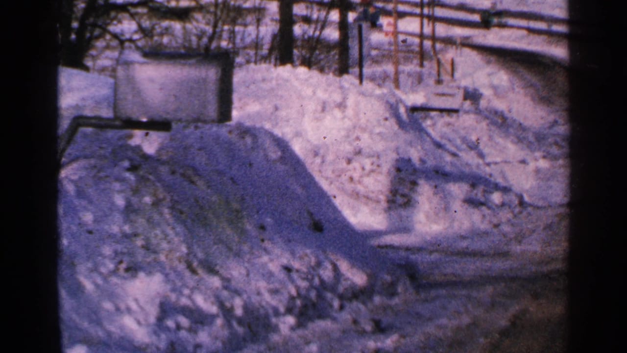Snow-covered Minnesota roadside in 1965, with a high drift, bare trees, utility lines, and soft blue-purple film color.