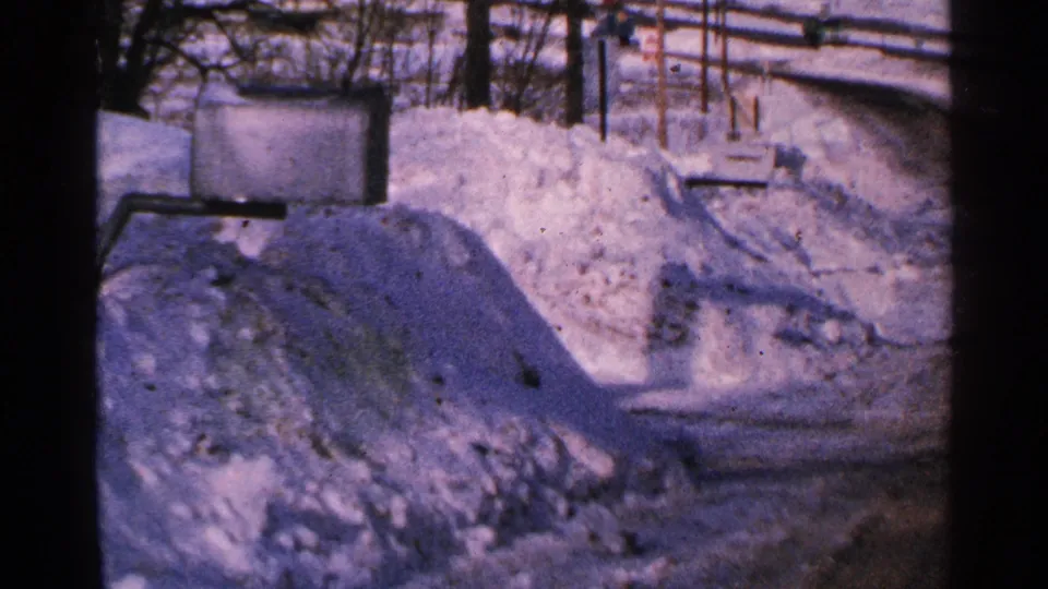 Snow-covered Minnesota roadside in 1965, with a high drift, bare trees, utility lines, and soft blue-purple film color.
