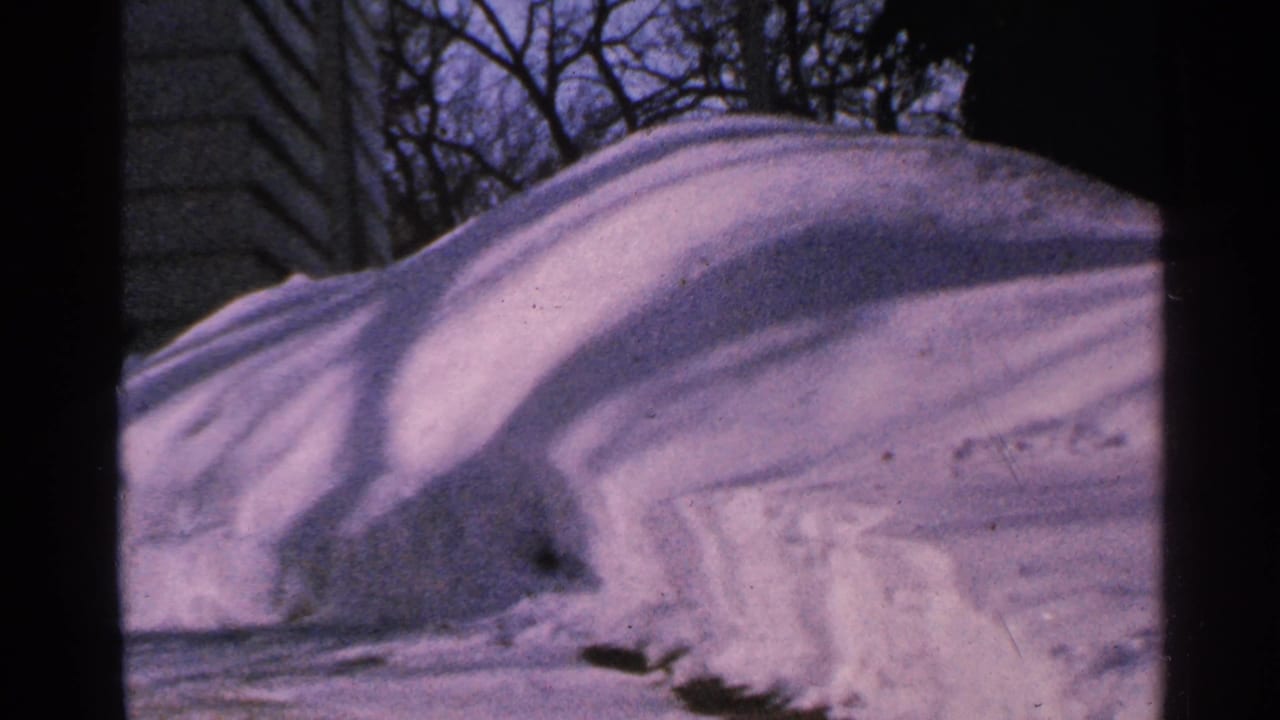 1965 Minnesota 8mm frame with a rounded snow drift, house siding, bare branches, and blue winter shadows.