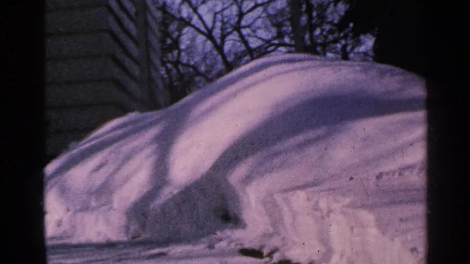 1965 Minnesota home-movie frame showing a high snowbank near siding, with leafless trees and shaded snow texture.