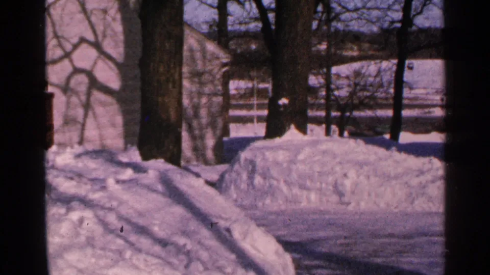 1965 Minnesota 8mm frame showing snow piled beside a cleared path, with trees, a building, and winter light beyond.