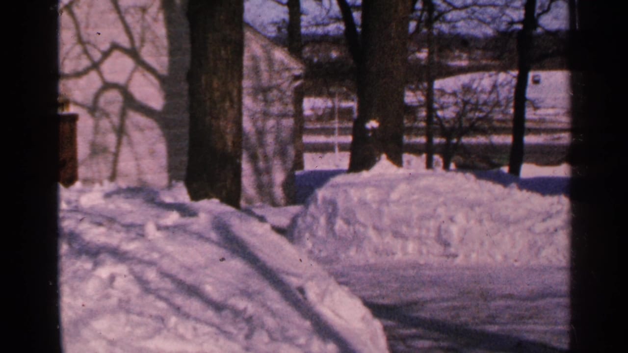 1965 Minnesota 8mm frame showing a snowy cabin-area yard with bare trees, piled snowbanks, and a pale building in winter light.