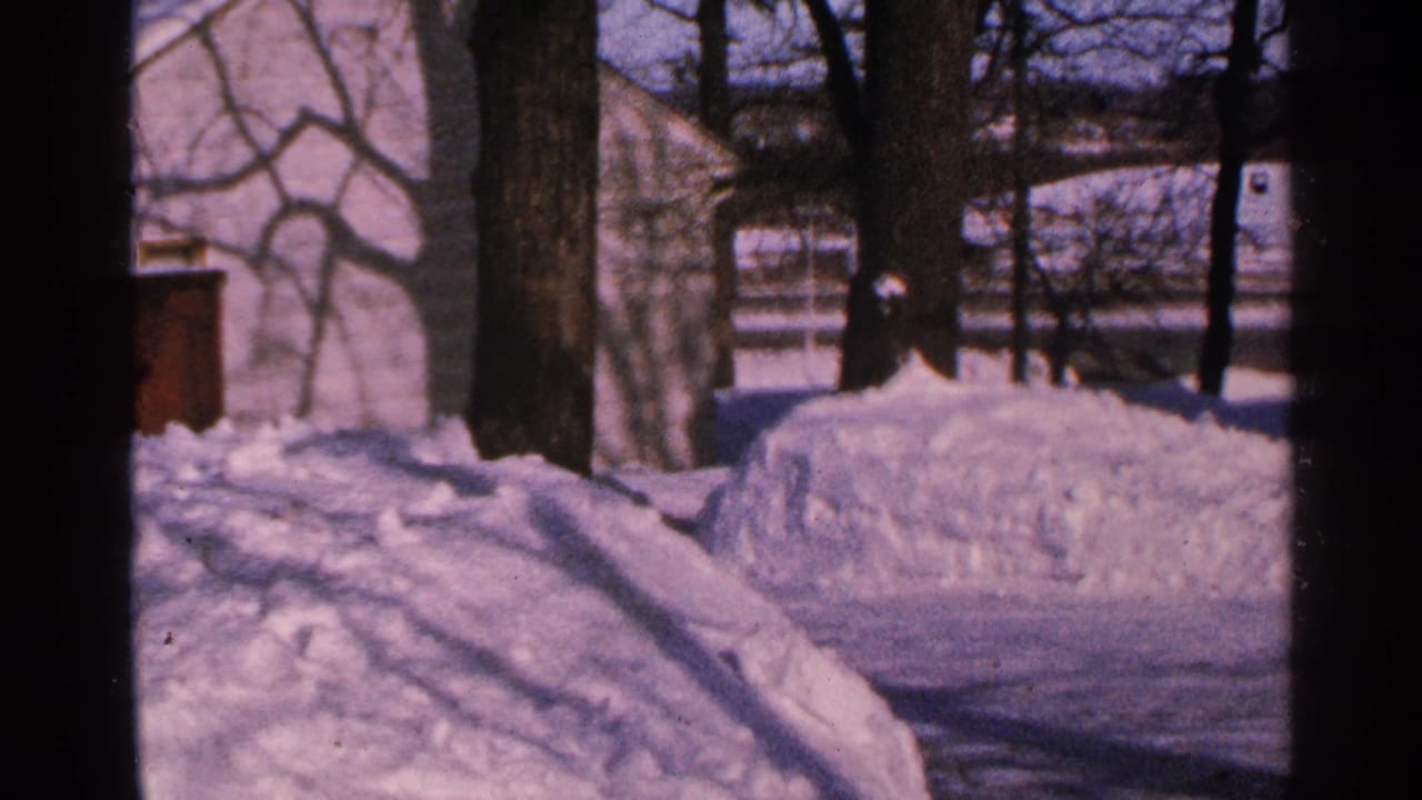 1965 Minnesota winter view of a snow-filled yard, bare tree trunks, and a light building partially obscured by small-gauge film framing.