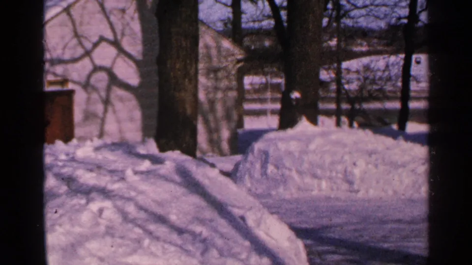 1965 Minnesota winter view of a snow-filled yard, bare tree trunks, and a light building partially obscured by small-gauge film framing.