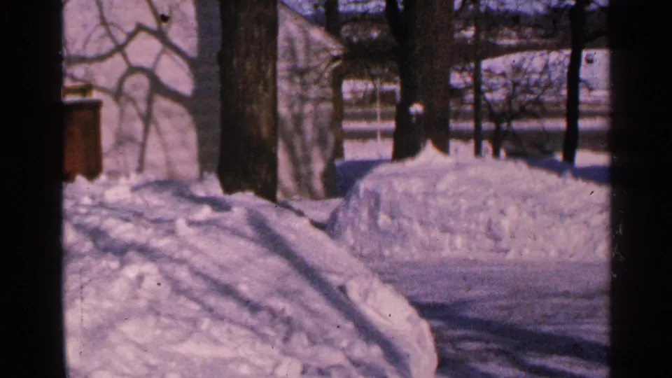 1965 Minnesota 8mm frame of a cabin-area winter yard, with long tree shadows crossing deep snow and a building in the background.