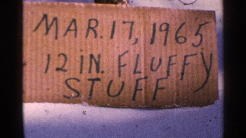Handwritten cardboard sign in snow reads Mar. 17, 1965 and 12 in. fluffy stuff, in color 8mm footage from Minnesota.
