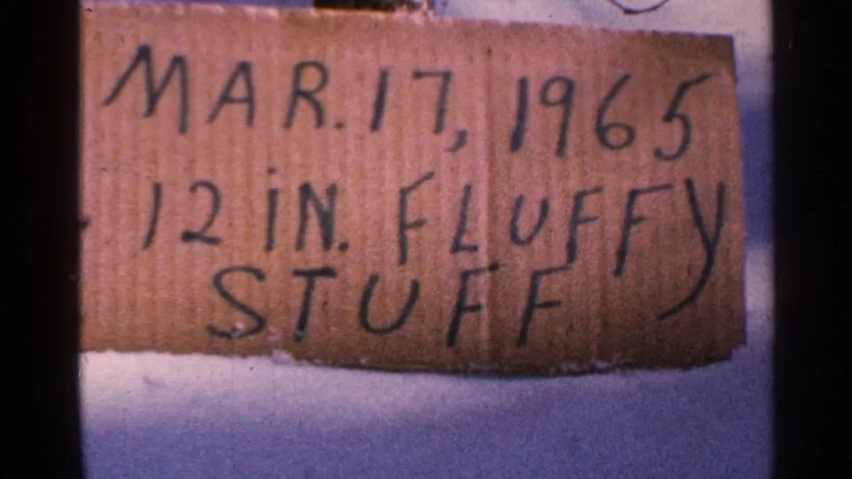Close-up of a handwritten cardboard sign on snow reading Mar. 17, 1965 and 12 in. fluffy stuff, with faded 8mm color.