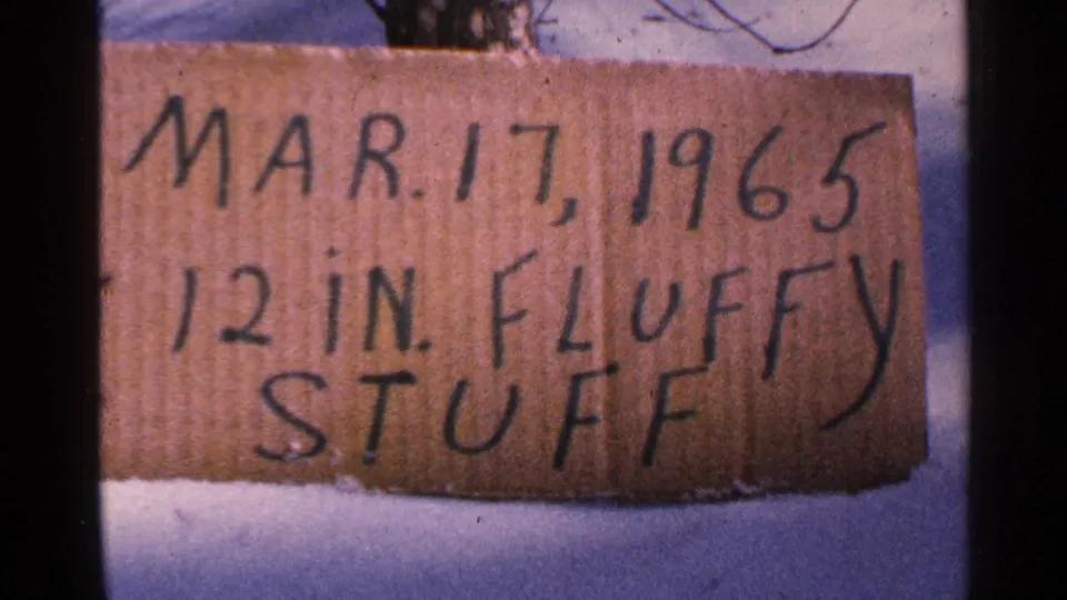 The 1965 Minnesota home-movie frame holds on the cardboard snowfall sign, with snow below and dark film borders at the sides.
