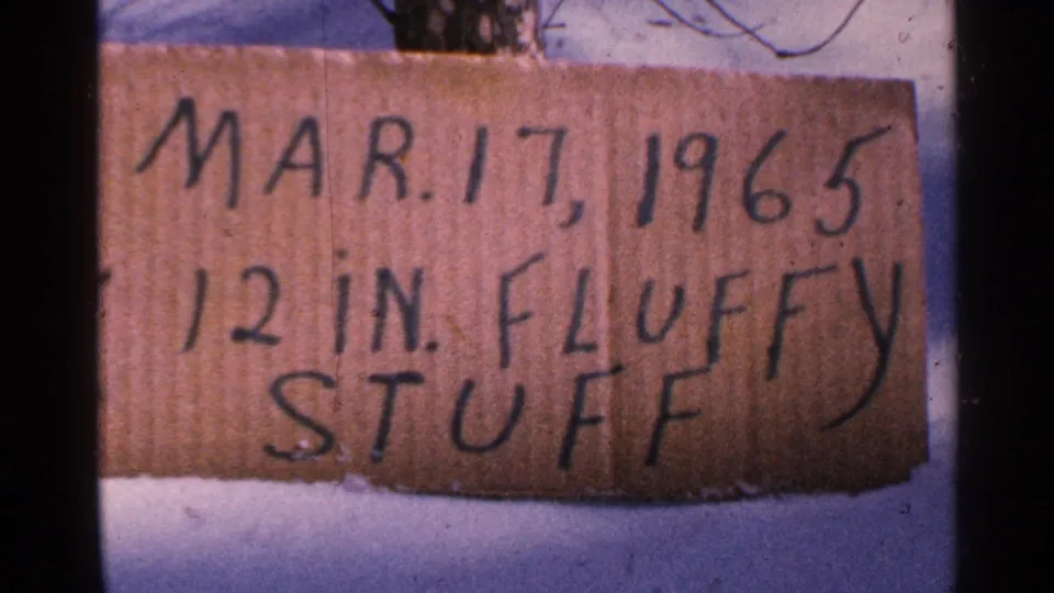 Minnesota 1965 8mm frame showing a handwritten cardboard snowfall sign set in snow, noting Mar. 17, 1965 and 12 inches.