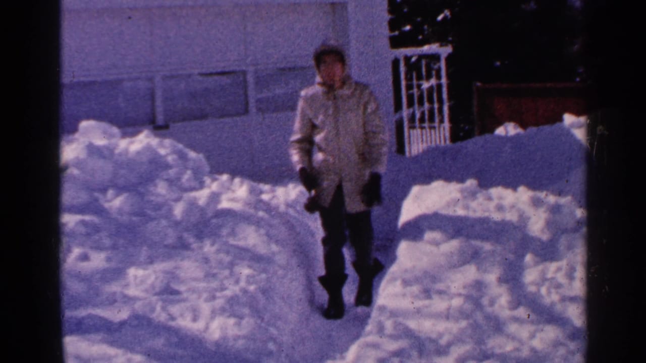 Minnesota 1965 home-movie frame of a woman in a light winter coat walking through deep snow beside a residential exterior.