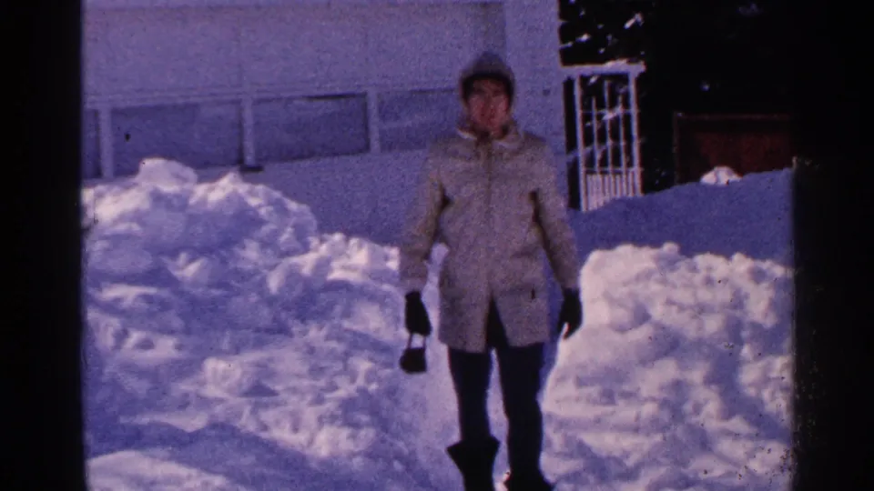 Minnesota 1965 8mm frame showing a woman closer to camera in a light winter coat, surrounded by piled residential snow.