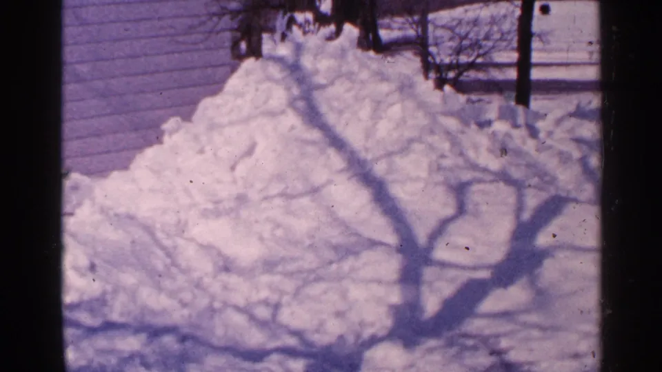 Minnesota 1965 home-movie frame of a high snowbank beside a residential wall, crossed by shadows from bare tree branches.