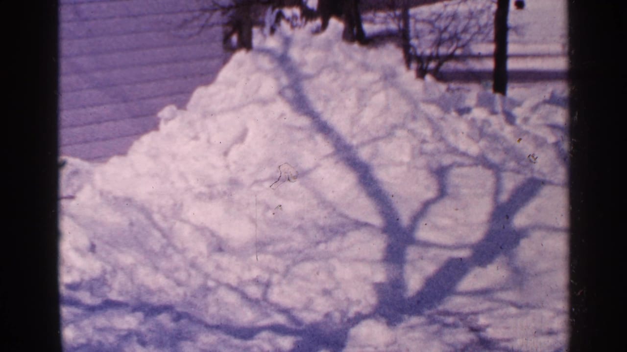 1965 Minnesota 8mm frame showing a large snow pile near a house exterior, crossed by the shadow of a bare tree.
