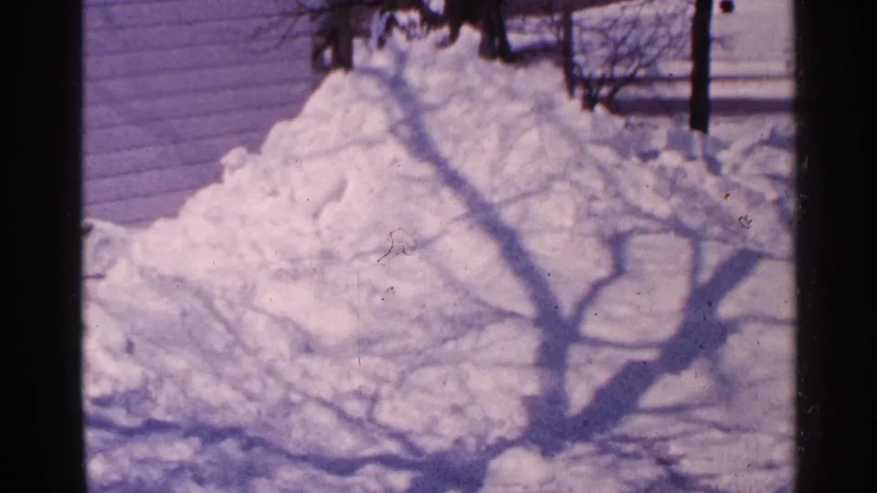 1965 Minnesota 8mm frame showing a large snow pile near a house exterior, crossed by the shadow of a bare tree.