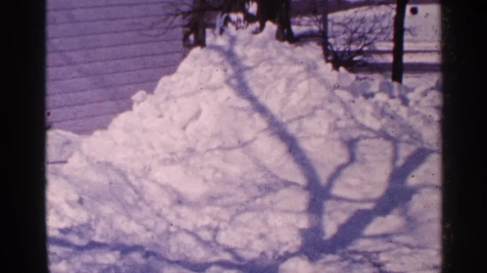 1965 Minnesota home-movie frame holding on a residential snow pile marked by bare tree shadows.