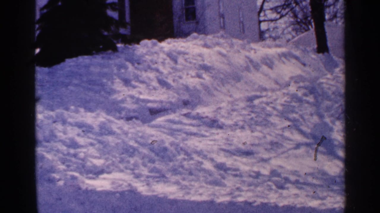 Tall snowbanks beside a driveway with a partial house and bare trees in the background.