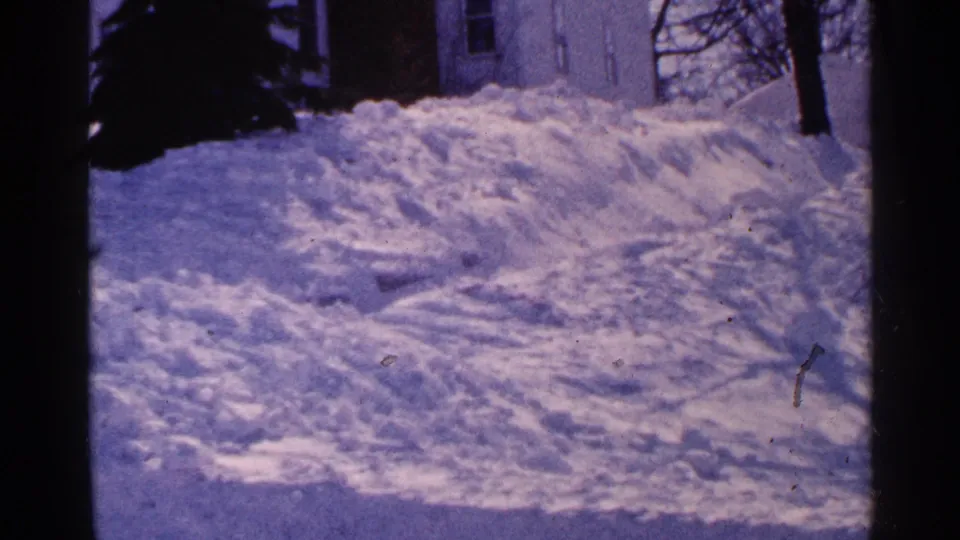 1965 Minnesota 8mm frame showing high plowed snowbanks near a residential driveway, with a partial house and bare winter trees behind them.